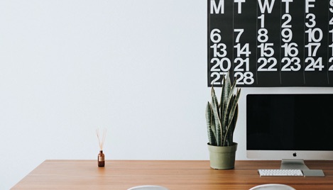 Wooden desk against a white wall with a computer monitor, plant, and air freshener on the desk.